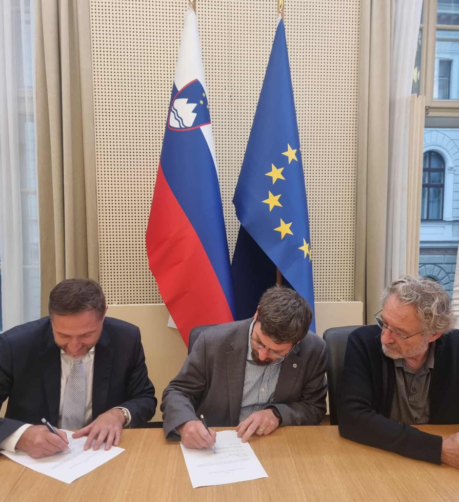 Podpisniki sedijo za omizjem in parafirajo dogovor, v ozadju slovenska in EU zastava