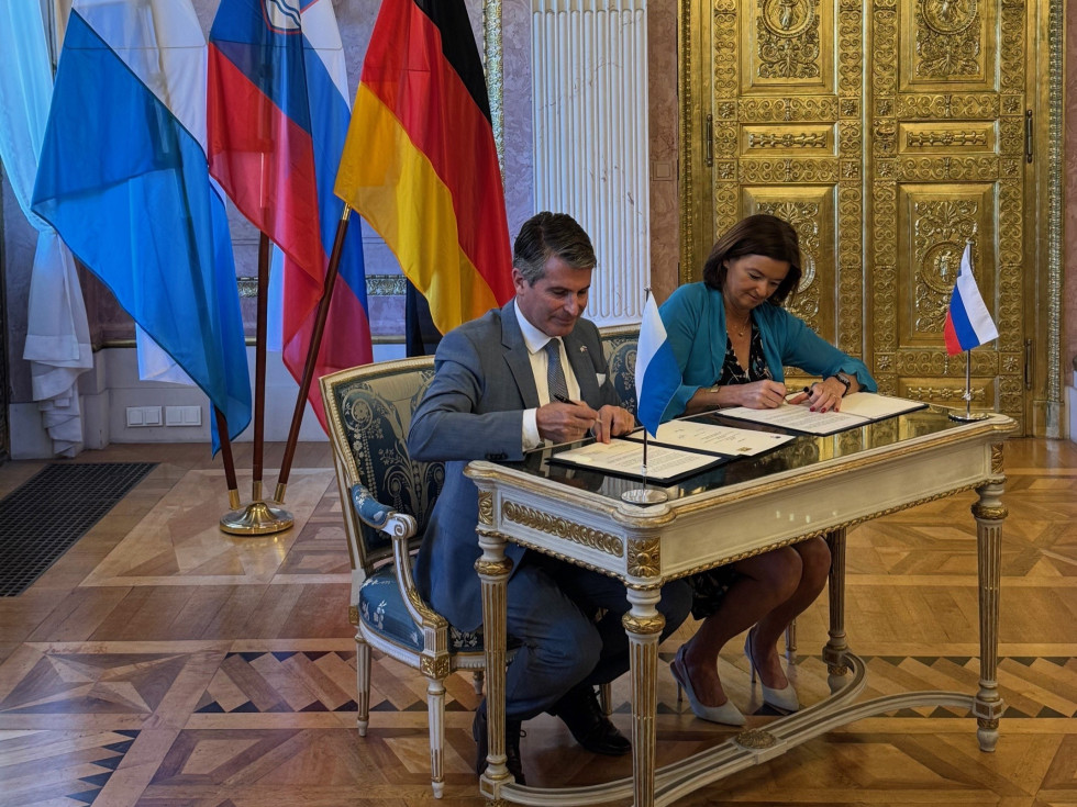 Ženska in moški sedita za mizo in podpisujeta vsak svoj dokument. Za njima so tri zastave.