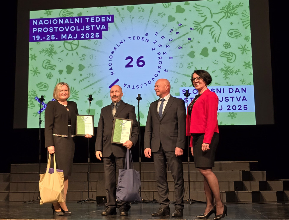 Štirje ljudje stojijo na odru, dva izmed njih v roki držita priznanja in vrečke. Vsi pozirajo fotografu.