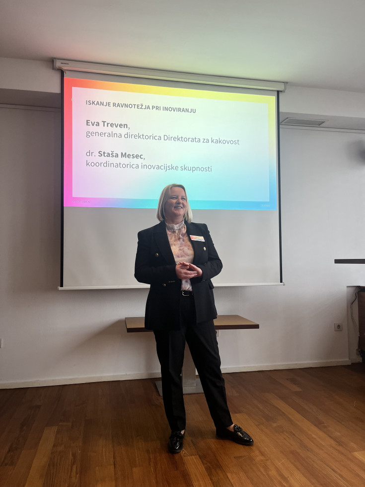 Predavateljica stoji v svetli sejni sobi in med predstavitvijo govori ob projekcijskem platnu, na katerem je prikazan diapozitiv z besedilom.