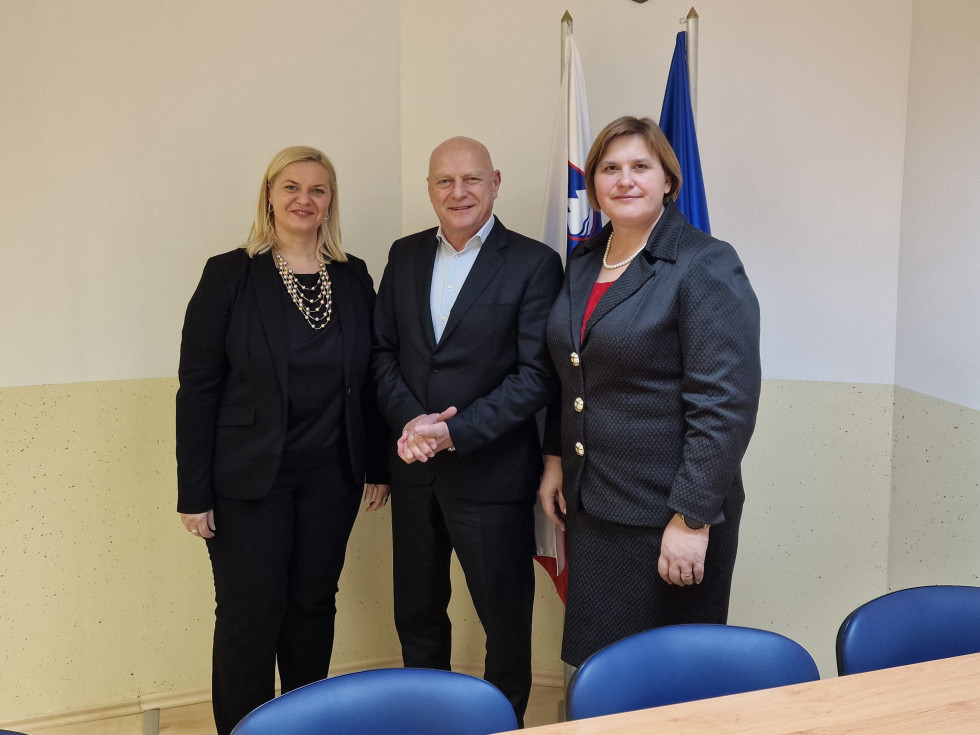 Trije ljudje stojijo, na sredini je moški, na vsaki strani sta ženski.