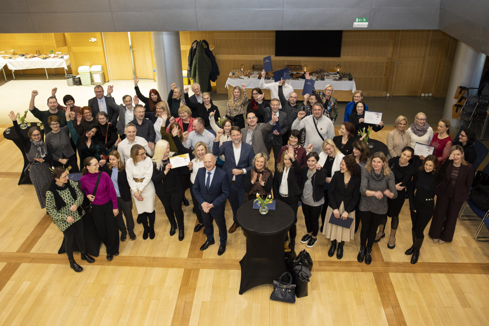 Skupinska fotografija udeležencev dogodka v dvorani; več deset ljudi stoji skupaj, nekateri dvigujejo roke in držijo priznanja.