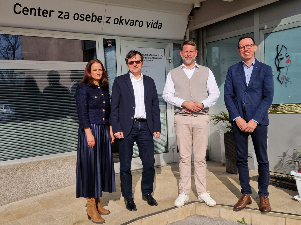 Štirje ljudje (ena ženska in trije moški) stojijo pred steklenimi vhodnimi vrati v stavbo in gledajo v objektiv.