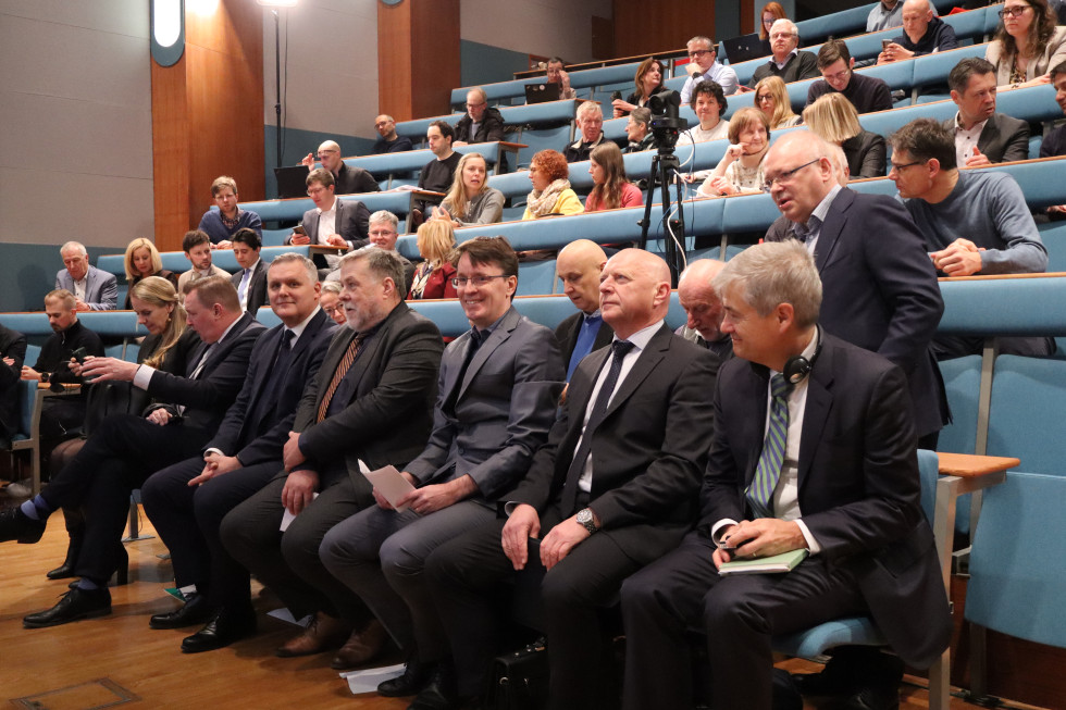 Gostje sedijo v dvorana in čakajo na začetek konference. Sedeži so postavljeni v nadstropjih (stopničasto)