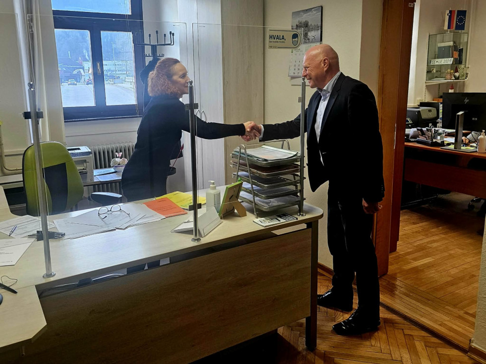 Minister se rokuje z uslužbenko v pisarni, med njima je zaščitna steklena pregrada.