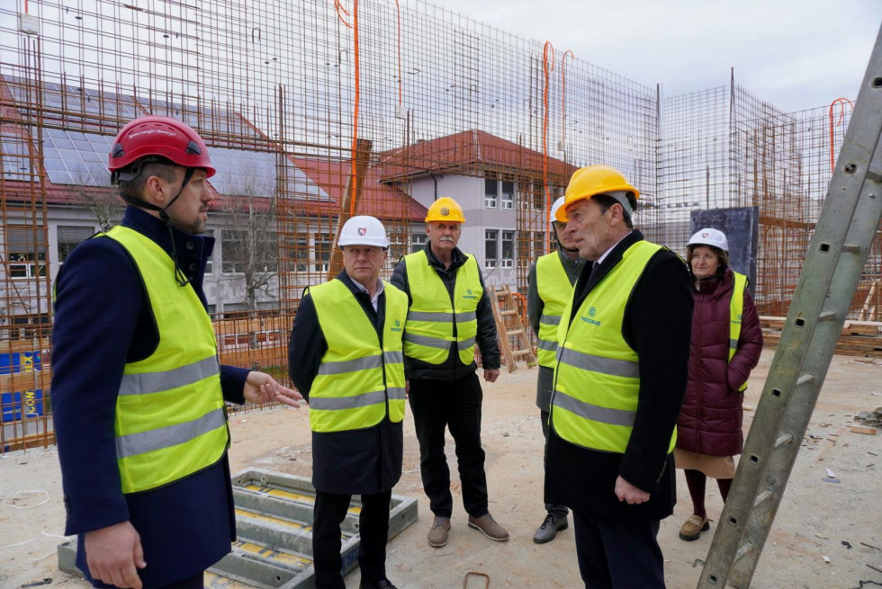 Minister mag. Franc Props in minister dr. Vinko Logaj z delegacijo med ogledom gradbišča novega vrtca v Šmartnem pri Litiji; udeleženci nosijo zaščitne čelade in odsevne jopiče, v ozadju potekajo armiranobetonska dela.