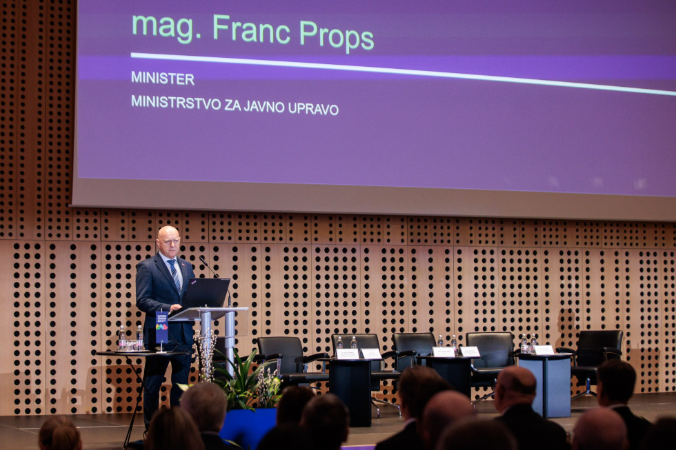 Moški stoji za govorniškim pultom na odru in govori.