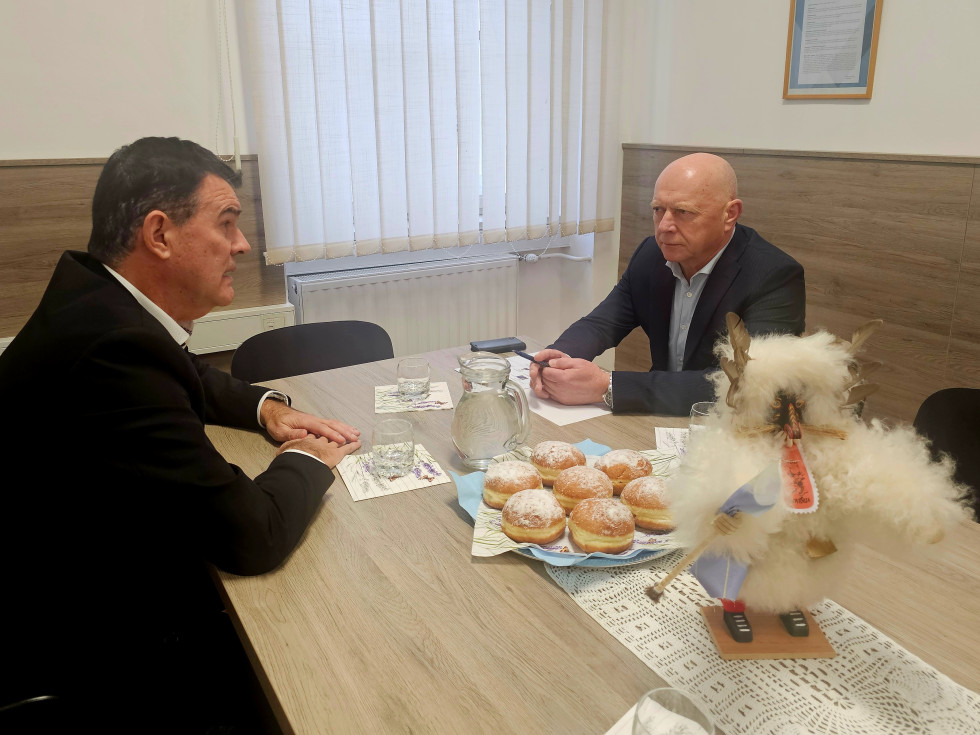 Načelnik upravne enote Ormož in minister za javno upravo (na desni) sedita za mizo v pisarni in se pogovarjata; na mizi vrč z vodo in kozarci, v ospredju pa stoji okrasna figura kurenta.