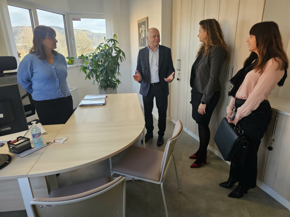 Minister v pisarni med pogovorom z zaposlenimi,; stojijo ob mizi in omarah, v ozadju je okno z razgledom na hribovito pokrajino.