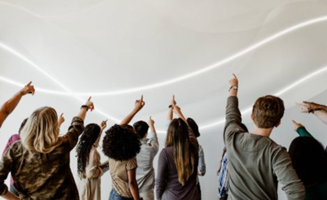 Skupina ljudi je postavljena s hrbti proti fotoaparatu. Gleda v zrak kamor tudi meri z eno roko in z iztegnjenim kazalcem.