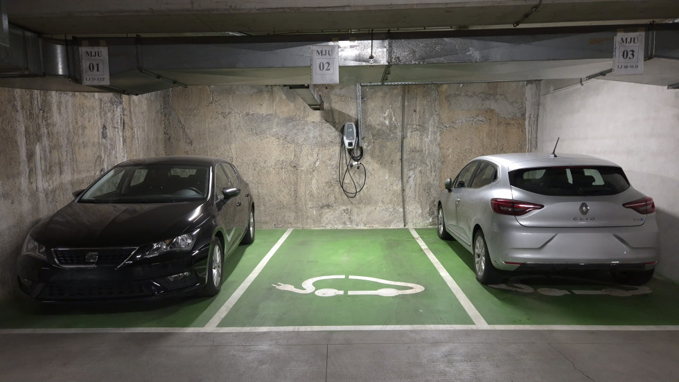 Tri parkirna mesta v garažni hiši. Na pravem in drugem sta parkirana avtomobila. Sredinsko parkirno mesto je prazno. Na steni pred njim je polnilnica za električna vozila. 