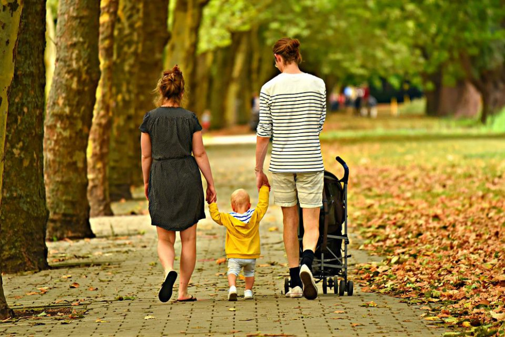 Starša, ki v parku sprehajata otroka. En otrok je v vozičku, en hodi.
