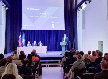 Na fotografiji za govornico stoji župan Mestne občine Koper, ki je gostiteljica posveta. V občinstvu sedijo vabljeni udeleženci posveta, to so županje in župani, direktorice in direktorji občinskih uprav ter vodje skupnih občinskih uprav..