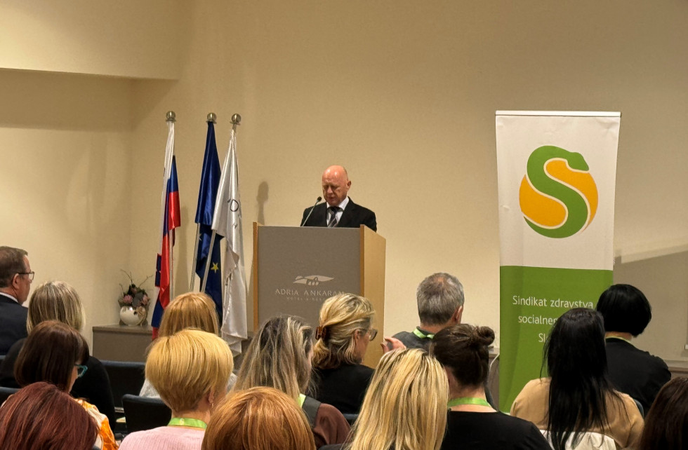 Od leve prosti desni so na fotografiji zastave, govornica, kjer udeležence seminarja pozdravlja minister za javno upravo, ter pano z napisom Sindikat zdravstva in socialnega varstva Slovenije