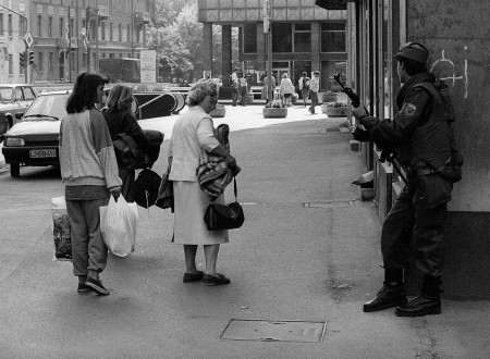 Pogled na ljubljansko ulico. Na levi strani pogled od zadaj na tri ženske, ki nosijo v rokah vreče in torbe. Na desni vojak s puško, ki je naslonjen na zid zgradbe, mimo katere gre skupina žensk.