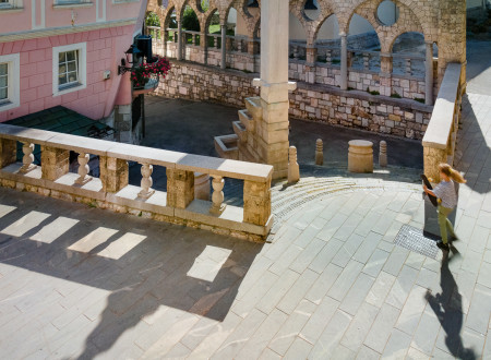 Roženvensko stopnišče v Kranju kot gledališka kulisa z arkadami v ozadju in petelinjim vodnjakom s terasasto postavljenimi kamnitimi posodami na sredini.