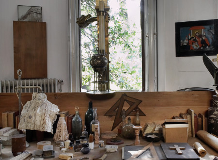 Delovna miza v Plečnikovi hiši, prekrita z raznovrstnimi predmeti: risalni pribor, ravnila, steklenice, sveča, lepilo, različni podstavki in knjige.
