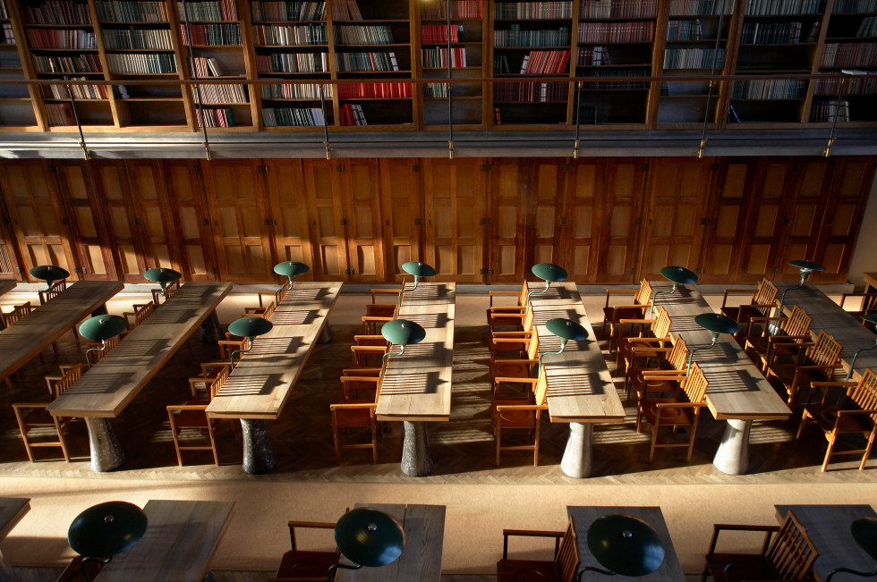 Prazni stoli in mize s svetilkami v čitalnici, v ozadju lesena stena, nad njo na galeriji knjižne police. 