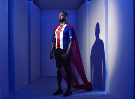 Temnopolti moški z desno nožno protezo oblečen v Supermana, s pokončno držo in odločnim pogledom navzgor proti levi