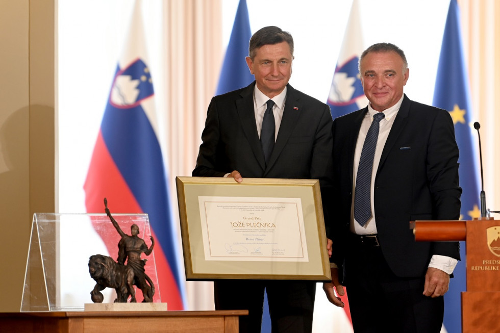 Portret dveh moških, s pogledom usmerjenim v kamero. Moški na levi drži v rokah uokvirjen zapis o prejemu nagrade, ki mu jo je vročil moški na njegovi levi