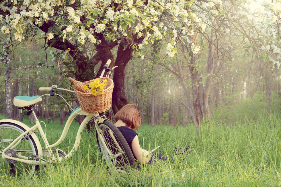 Dekle, ki sedi v travi ob cvetočem drevesu, je s hrbtom naslonjena na kolo in bere knjigo