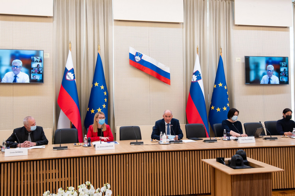 Predsednik vlade in člani sveta za konferenčnim omizjem, v ozadju zastave. 