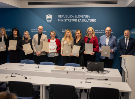 Predstavniki in predstavnice občin stojijo drug ob drugem in v roki držijo razprto mapo s priznanjem, za njimi na modrem platnu logotip Ministrstva za kulturo