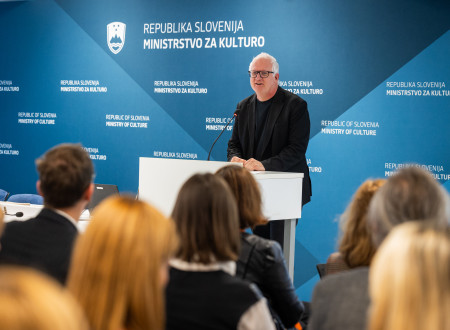 Državni sekretar stoji za belim govornim pultom, pred njim sedijo udeleženci dogodka 