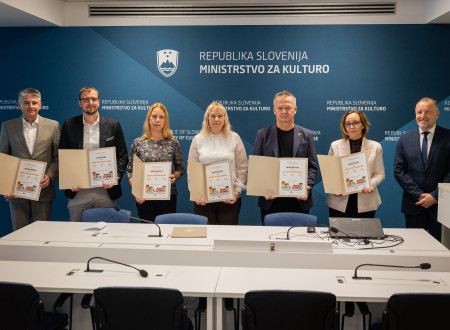 Predstavniki in predstavnici občin prejemnic stojijo drug ob drugem in v roki držijo razprto mapo s priznanjem, za njimi na modrem platnu logotip Ministrstva za kulturo