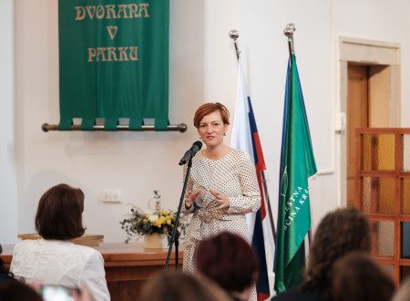 Ministrica za kulturo stoji pred mikrofonom, pred njo sedi nekaj ljudi, za njo slovenska zastava in zastava Mestne občine Krško 