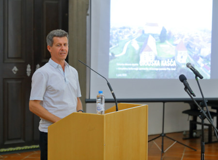 Državni sekretar stoji za govorniškim odrom z mikrofonom in nagovarja zbrane
