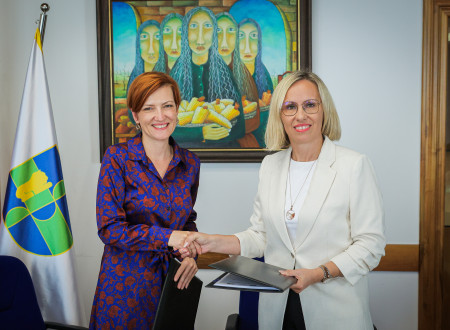 Ministrica za kulturo in županja sta nasmejani in se rokujeta. V rokah držita modro mapo s podpisanimi izvodi pogodbe