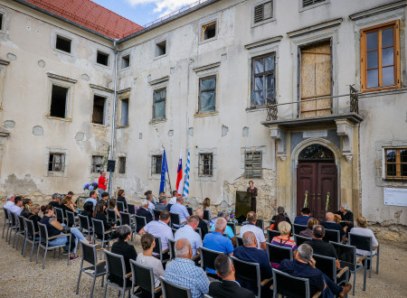 Udeleženci odprtja dvorca sedijo na stolih, razporejenih v vrste na dvorišču dvorca, in poslušajo govor ministrice za kulturo, ki stoji za govorniškim odrom pred vhodnimi vrati v dvorec