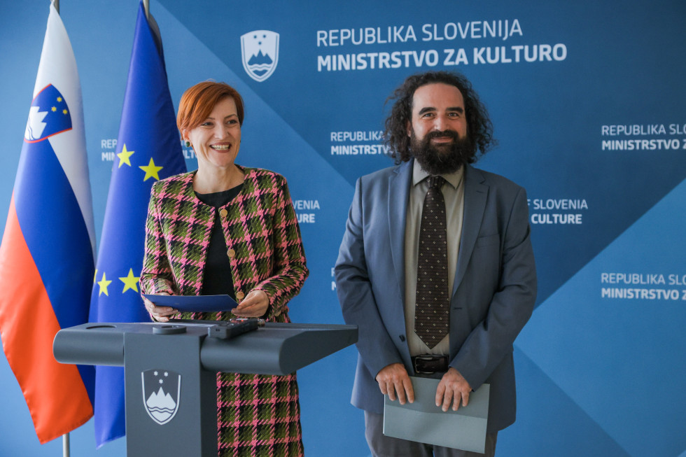 Nasmejana ministrica za kulturo in generalni direktor stojita drug ob drugem, za njima modro ozadje, na katerem je logotip Ministrstva za kulturo, na desni strani ministrice zastava Evropske unije in slovenska zastava