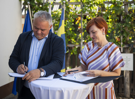 Župan in ministrica za kulturo stoje podpisujeta pogodbo za visoko barsko mizo, pokrito z belim prtom