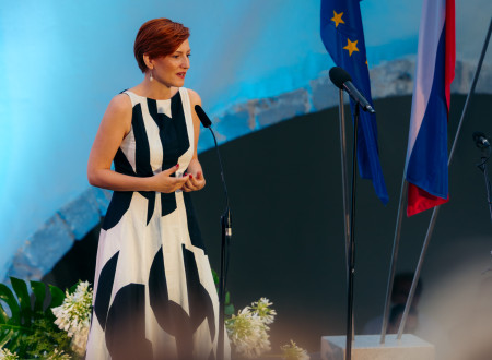 Ministrica za kulturo stoji pred mikrofonom. Na njeni levi strani zastavi Evropske unije in Slovenije