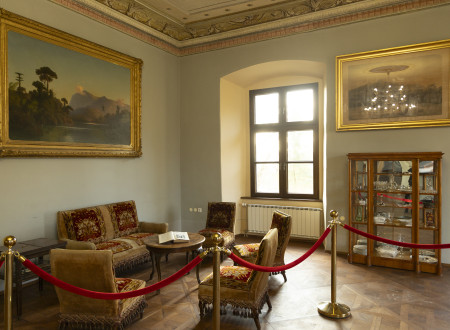  Z vrvicami zavarovani oblazinjeni stoli, podobni foteljem, postavljenimi okrog nizke lesene mize, levo veliko okno, ki osvetljuje prostor, desno in levo od okna na steni visita sliki, na levi pod sliko stoji steklena vitrina