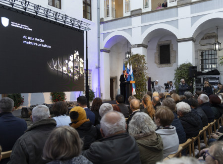 Ministrica za kulturo stoji za govorniškim pultom na odru in nagovarja zbrane, ki na stolih sedijo pod odrom