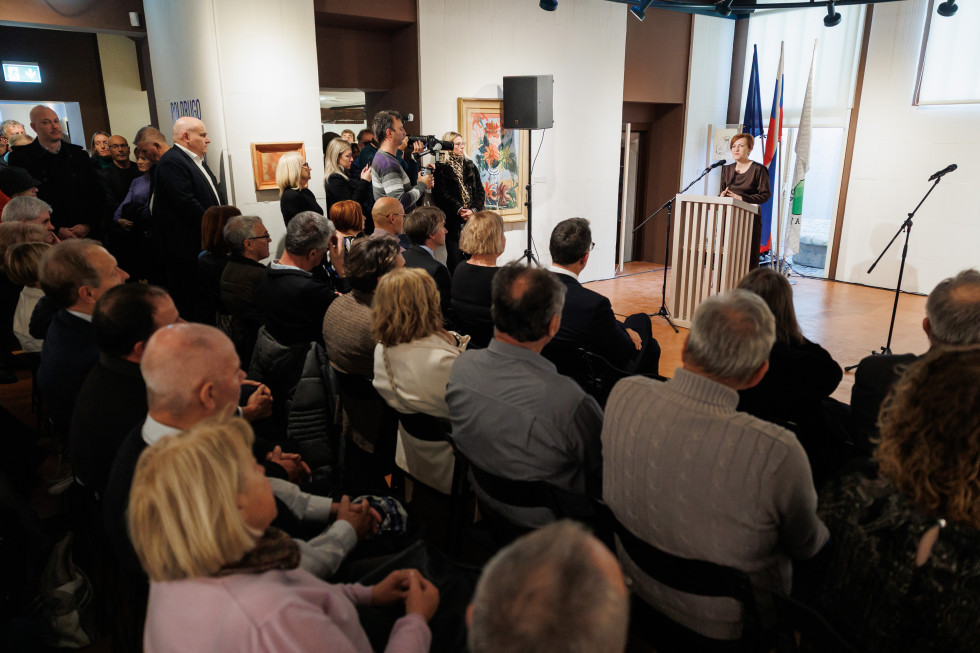 Ministrica za kulturo stoji za govornim pultom, pred njo poslušalci sedijo in stojijo