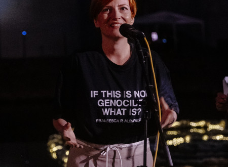 Ministrica pred mikrofonom nagovarja zbrane na večernem festivalskem dogdku, na njeni črni majici napis If this is not genocide, what is it? 