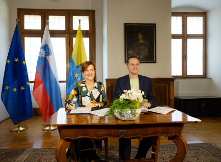 Nasmejana ministrica za kulturo in župan Celja sedita rjavo leseno masivno mizo, na sredini katere je bel rožni aranžma