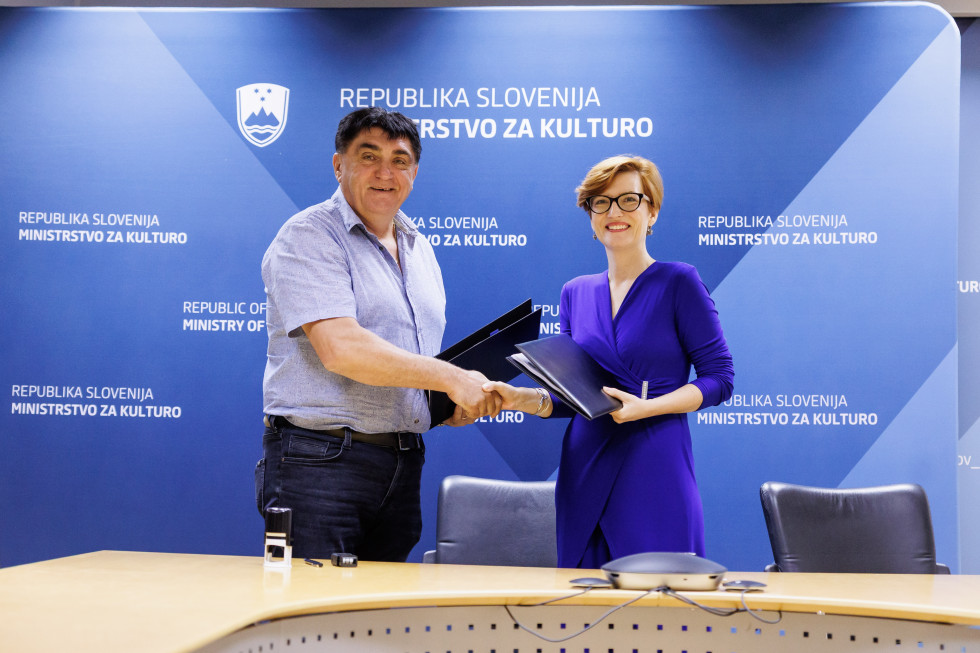 Župan in ministrica se rokujeta, v levi roki držita modro mapo, v kateri je podpisana pogodba