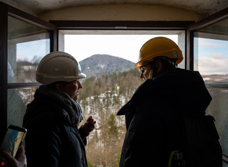 Ministrica za kulturo še z drugo osebo gleda skozi okno, pogled na poraščene in delno zasnežene griče