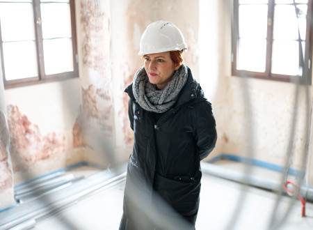 Ministrica za kulturo s čelado na glavi, s šalom in v bundi, stoji v svetlem prostoru, ki se prenavlja