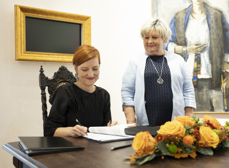 Ministrica za kulturo sedi za mizo in podpisuje pogodbo, na njeni levi strani stoji županja