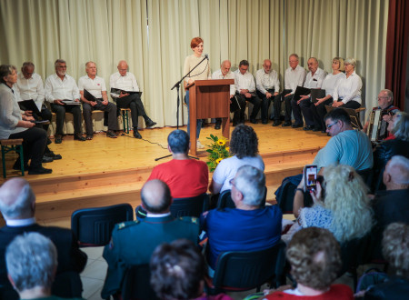 Ministrica za kulturo stoji na odru za govorniškim pultom in nagovarja zbrane. Za njo ob steni sedijo pevci, oblečeni v belo srajco in črne hlače, pod odrom sedijo udeleženci slovesnosti