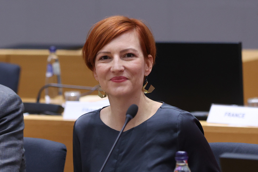 Ministrica za kulturo z nasmehom sedi za eno od miz, ki so razporejene v krogu in v več vrstah. Pred njo mikrofon.