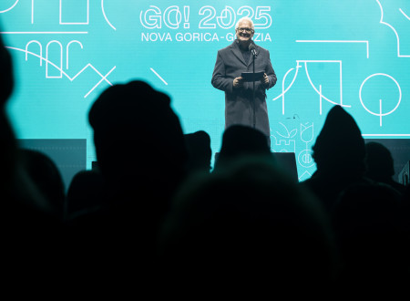 Državni sekretar, oblečen v plašč, stoji na odru pred mikrofonom, za niim modrozeleno ozadje evropske prestolnice kulture