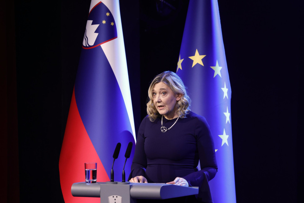 Ministrica stoji za govorniškim odrom, za njo slovenska zastava in zastava Evropske unije