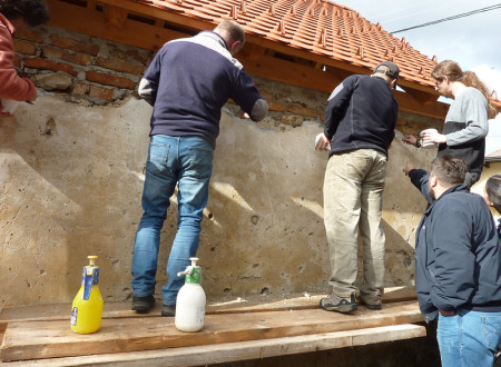 Štirje moški stojijo na lesenem odru ob hiši in ročno obnavljajo fasadni omet